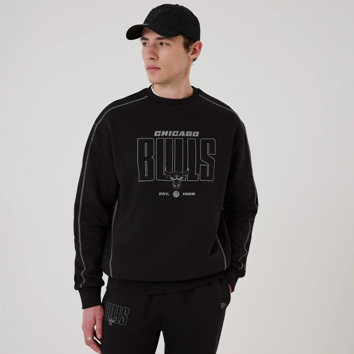 New Era Chicago Bulls NBA Graphic Black Crew Neck Sweater, front view on a model wearing a matching black cap, featuring Bulls lettering and logo across the chest, accented with grey contrast stitching along the sleeves.