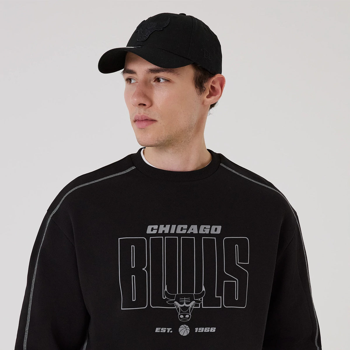 New Era Chicago Bulls NBA Graphic Black Crew Neck Sweater, front view on a model wearing a black cap, featuring grey Bulls lettering across the chest with the team's bull logo and Est. 1966 printed below.