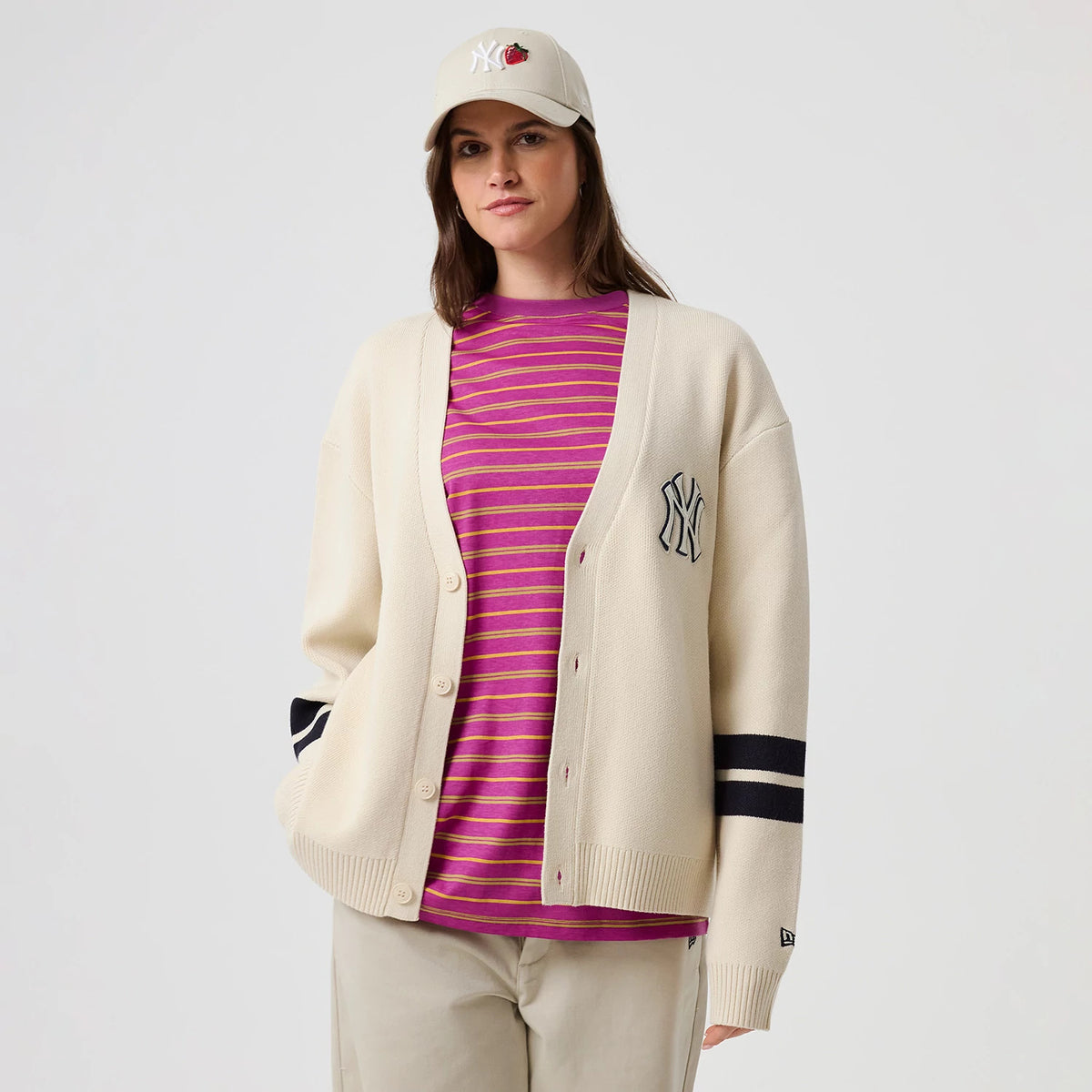 New Era New York Yankees MLB Knit Light Beige Cardigan, front view on a model with a striped pink and purple underlayer, showing an embroidered NY logo on the left chest, beige buttons, and black and white striped on the right sleeve.