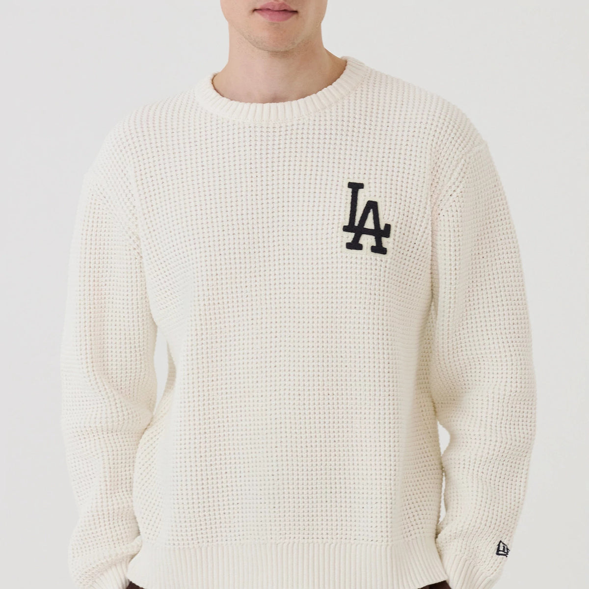 New Era LA Dodgers MLB Waffle Knit Open White Crew Neck Sweater, front view on a model, featuring a prominent black LA logo on the left chest, a textured waffle knit pattern, and the New Era flag logo on the left sleeve cuff.