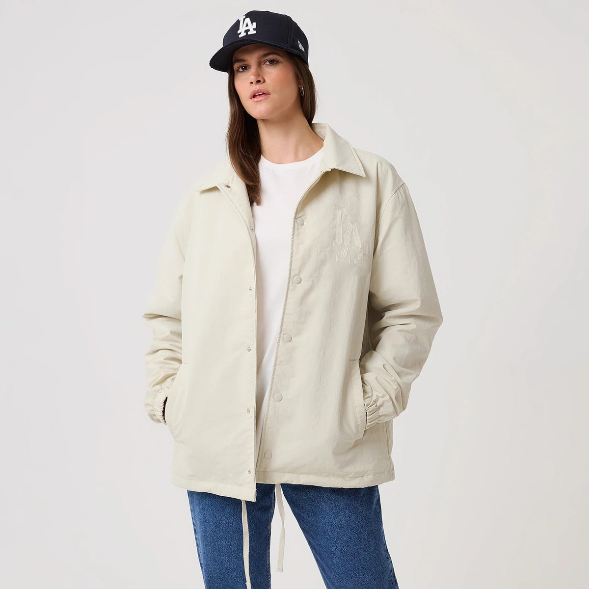 New Era LA Dodgers MLB Light Beige Coach Jacket, front view on a model styled with a dark cap, displaying the embroidered LA Dodgers logo on the left chest, button-down closure through the beige fabric, and sleeves gathered at the wrists.