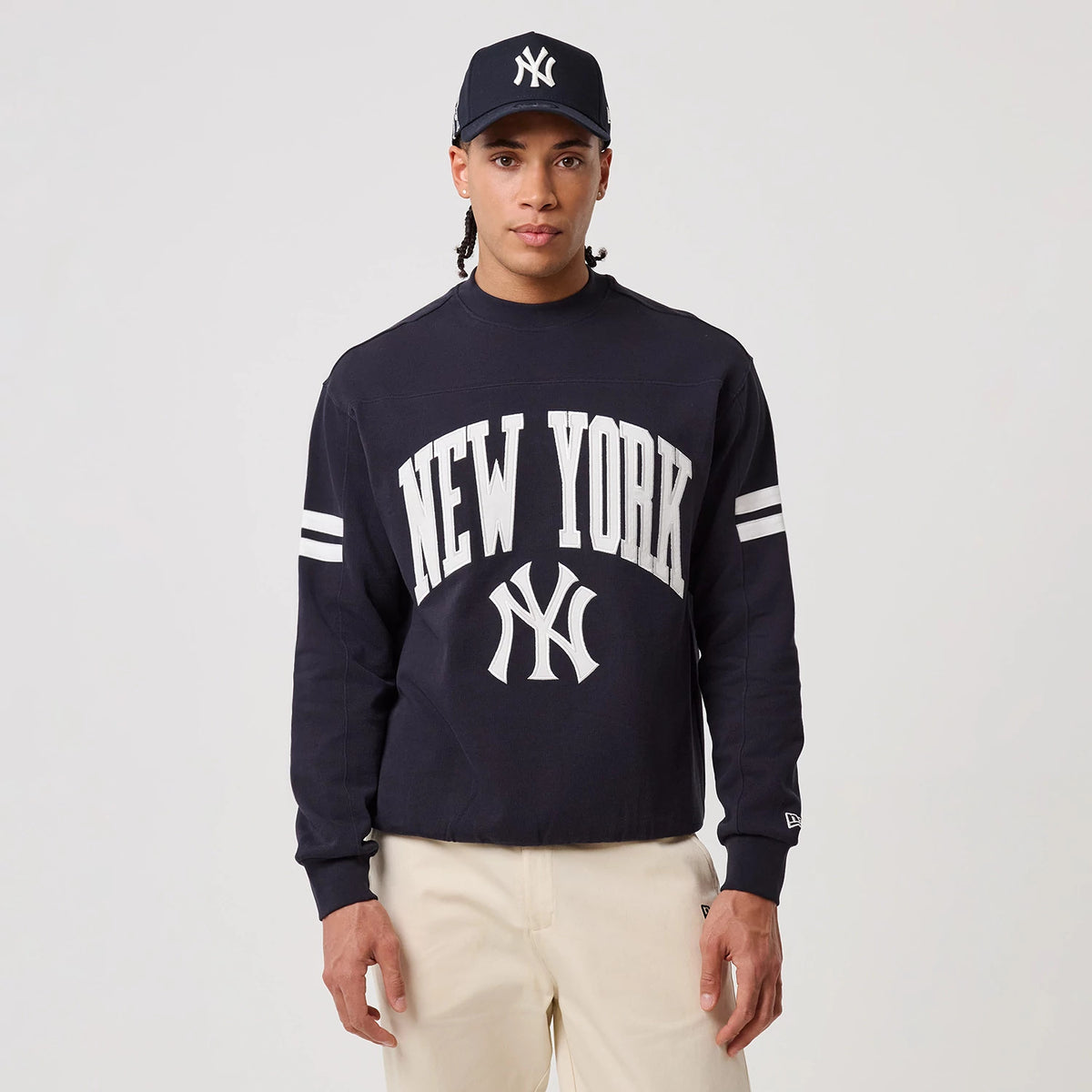 New Era New York Yankees MLB Arch Graphic Navy Crew Neck Sweater, front view on a model wearing a matching cap, featuring white New York lettering across the chest, the Yankees logo, and dual white stripes on each sleeve.