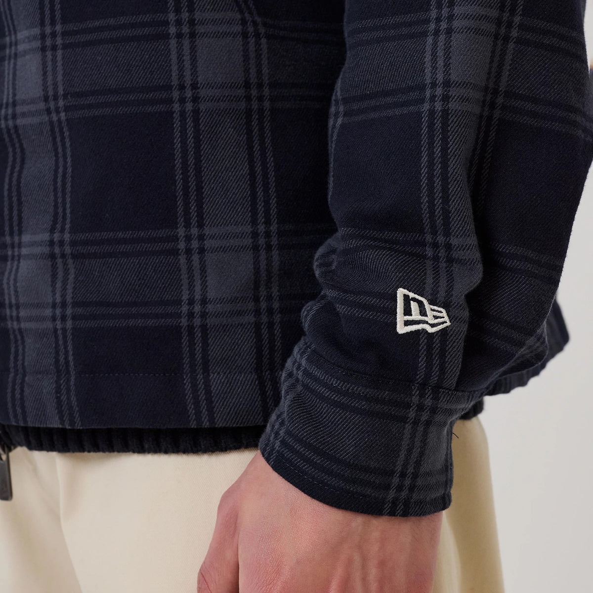 New Era Navy Shacket, close-up view of a sleeve showing dark navy and grey chequered pattern, embroidered white New Era flag logo on the left sleeve, and ribbed elastic hem above beige trousers.