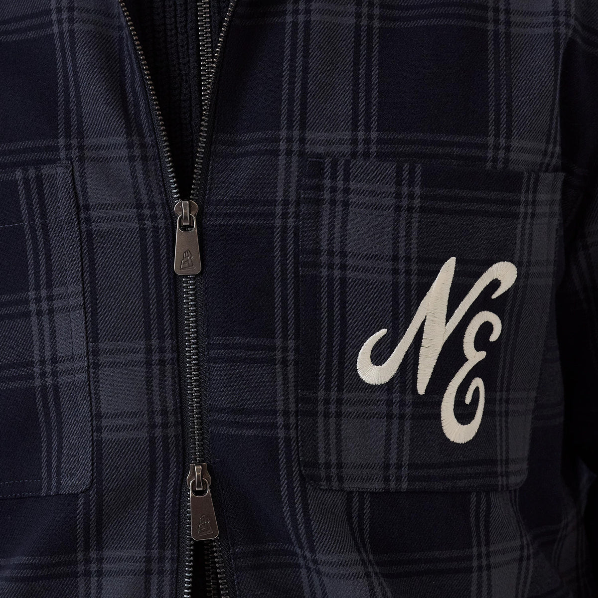 New Era Navy Shacket, front view showing a navy plaid pattern with twin chest pockets, each featuring a zip closure, and a white embroidered NE on the right pocket, highlighting the shacket's casual and versatile style.