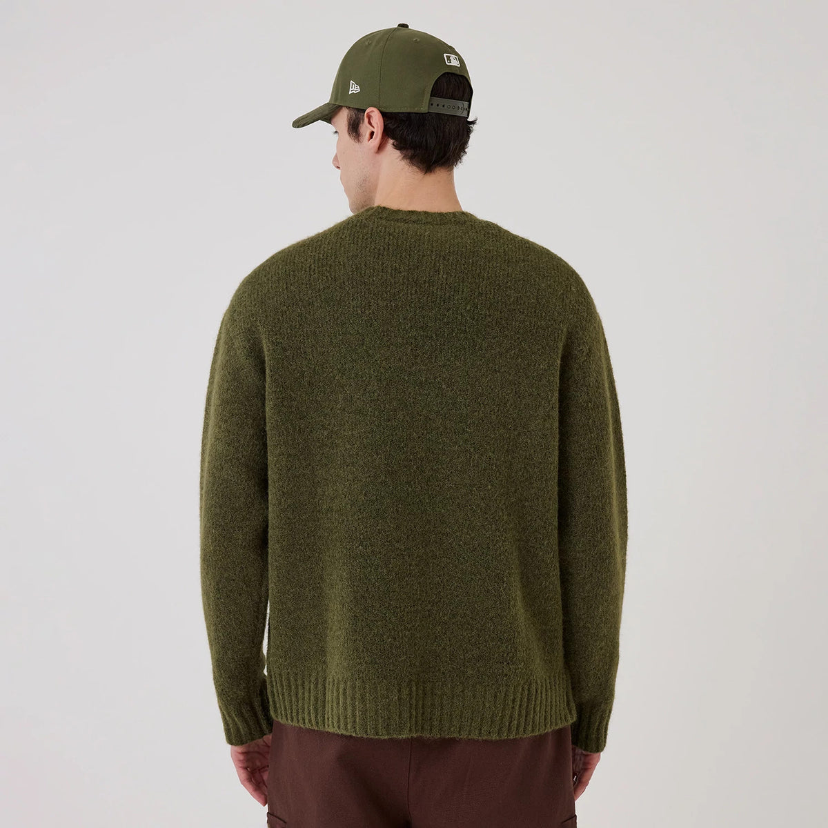New Era Simple Knit Green Knitted Sweater, back view on a model wearing a matching green cap and brown trousers, showing a relaxed fit with a ribbed hem and cuffs, in a textured green knit fabric.