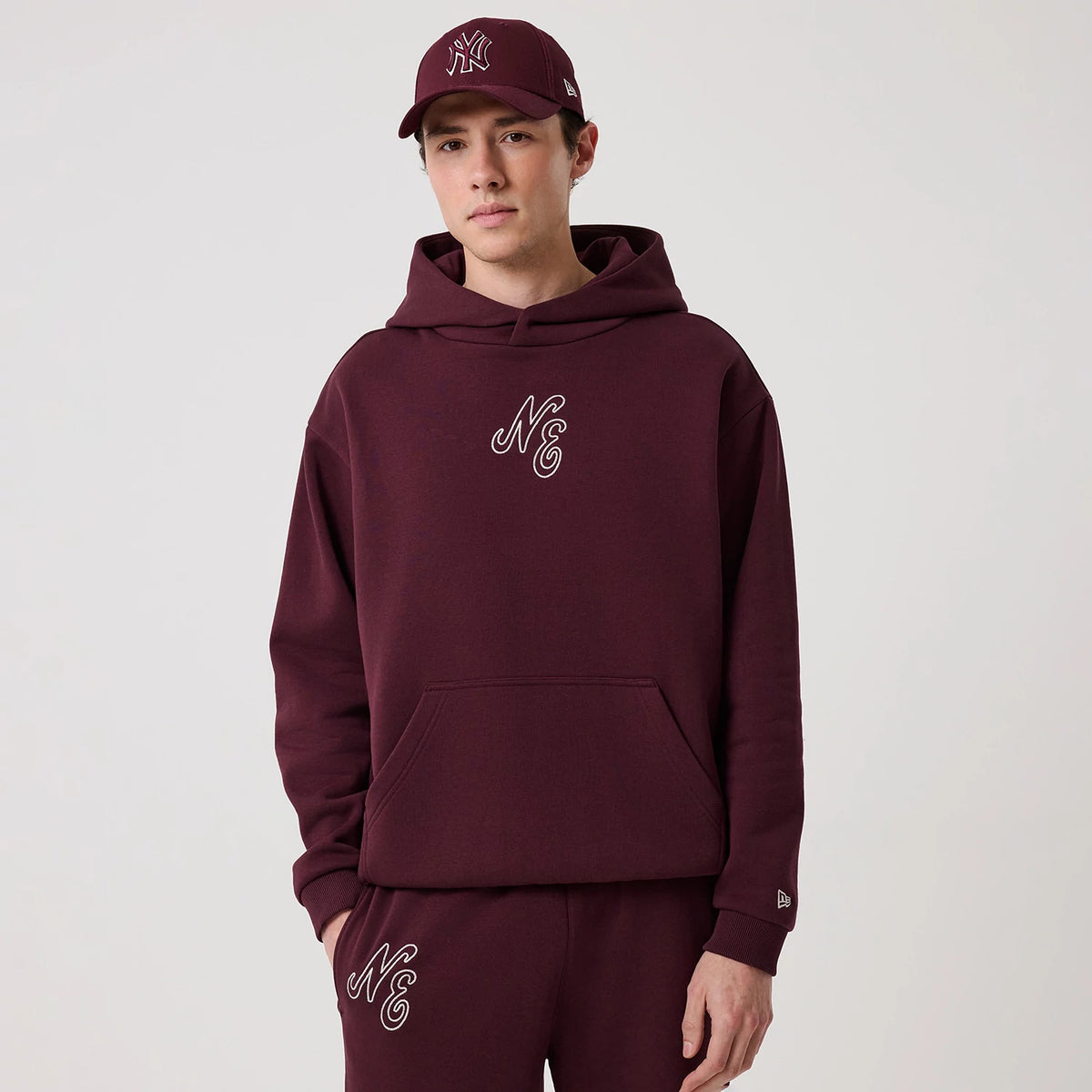 New Era Crest Graphic Dark Purple Pullover Hoodie, front view on-model showing an embroidered NE logo on the chest, a large kangaroo pocket at the front, and a small embroidered New Era logo on the left sleeve cuff.