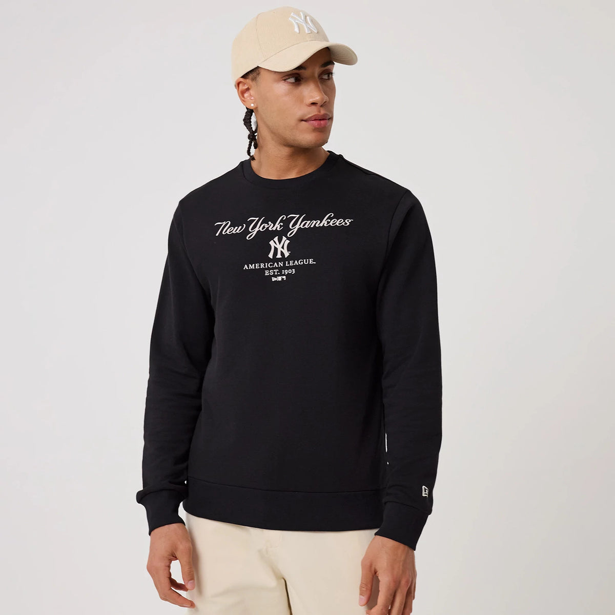 New Era New York Yankees MLB Linear Black Crew Neck Sweater, on-model front view featuring a white New York Yankees script and logo on the chest, paired with a beige cap, and a small New Era flag logo on the left sleeve cuff.