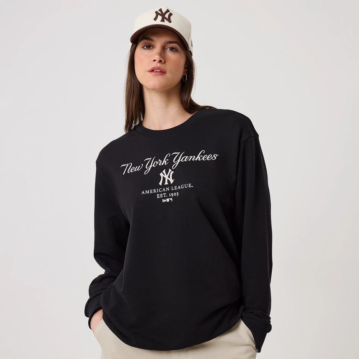 New Era New York Yankees MLB Linear Black Crew Neck Sweater, on-model front view with the New York Yankees script and logo embroidered in white on the chest, featuring long sleeves and a relaxed fit paired with a white cap.