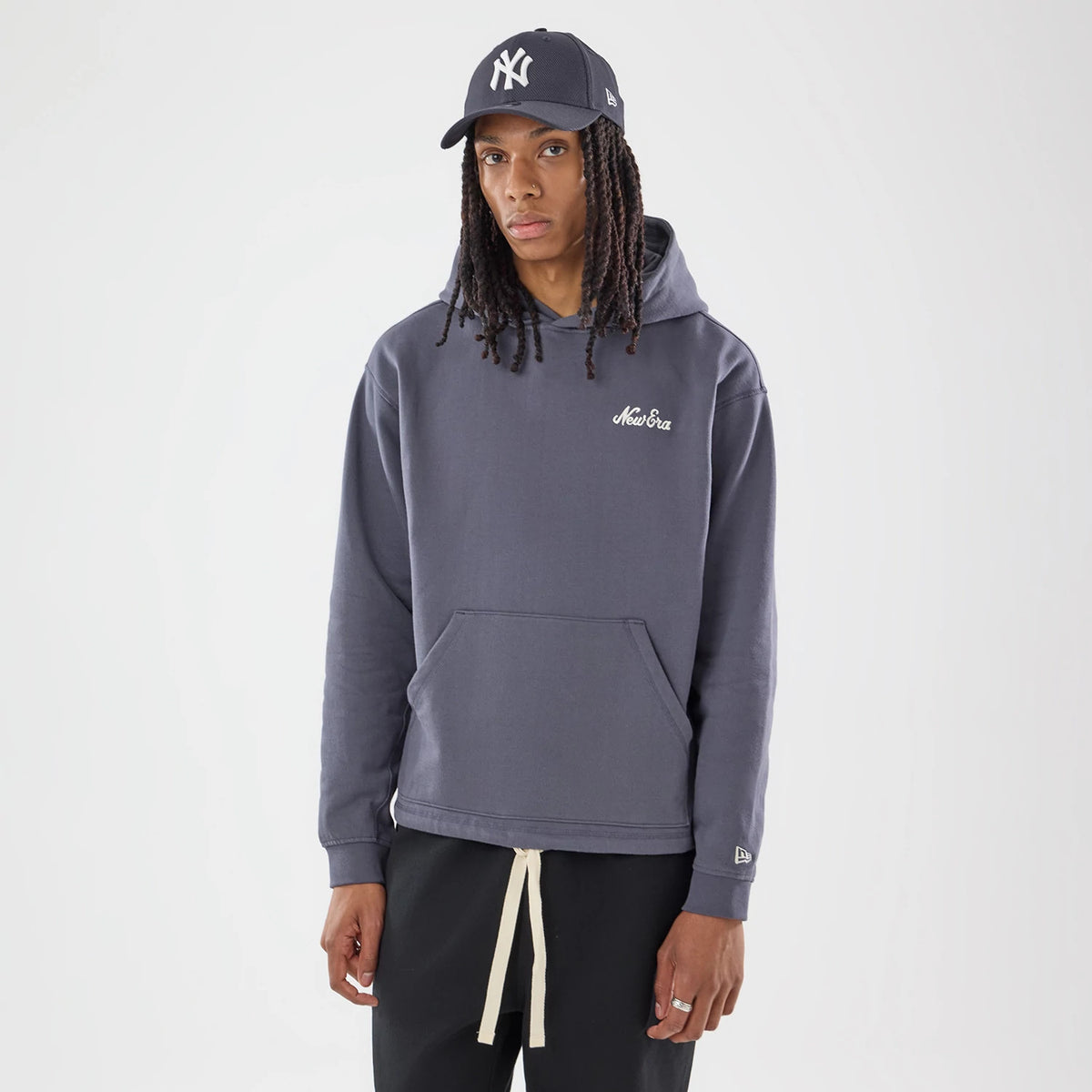 New Era Open Hem Grey Pullover Hoodie, front view on a model wearing a matching cap, showcasing an embroidered New Era logo on the chest, a large kangaroo pocket, and a sewn New Era flag logo on the lower left sleeve.