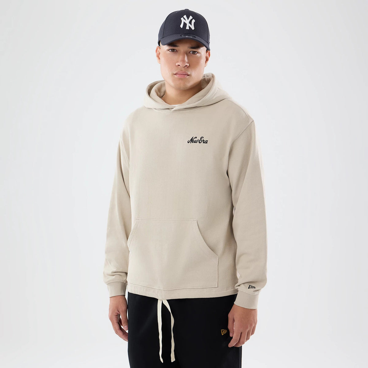 New Era Open Hem Light Beige Pullover Hoodie, front view on-model displaying the light beige colour with a kangaroo pocket, New Era embroidered logo on the chest, and a small embroidered logo on the left sleeve cuff.