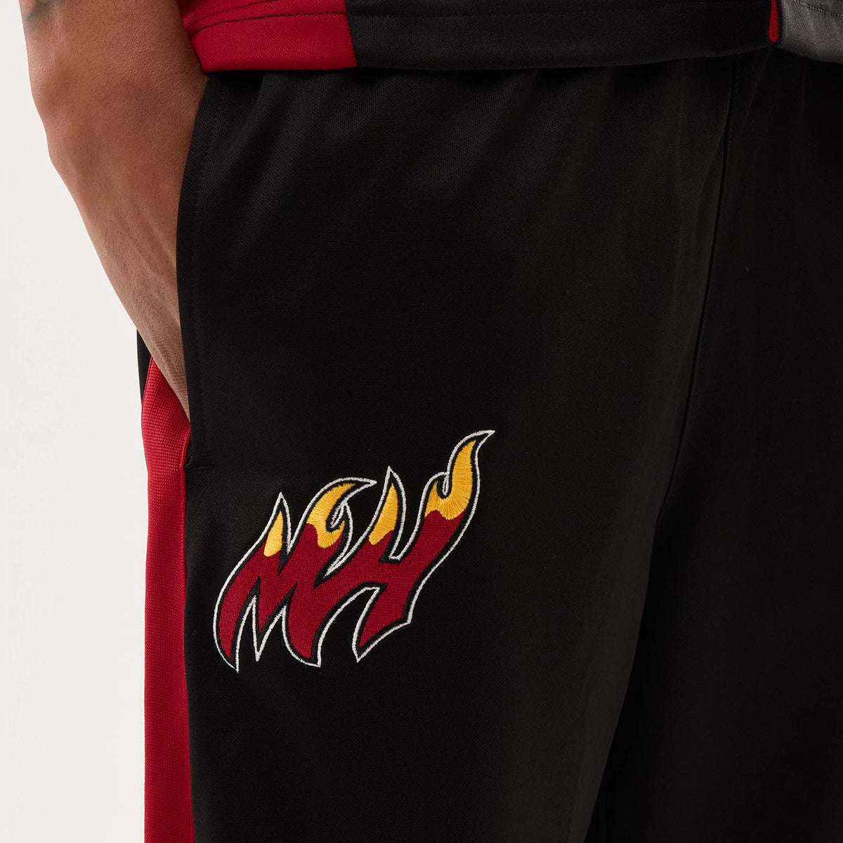 New Era Miami Heat NBA Hardwood Classic Straight Black Straight Leg Joggers, close-up view showing the flaming MH logo embroidered in red, yellow, and white on the left leg, with contrasting red side panels next to the pocket opening.