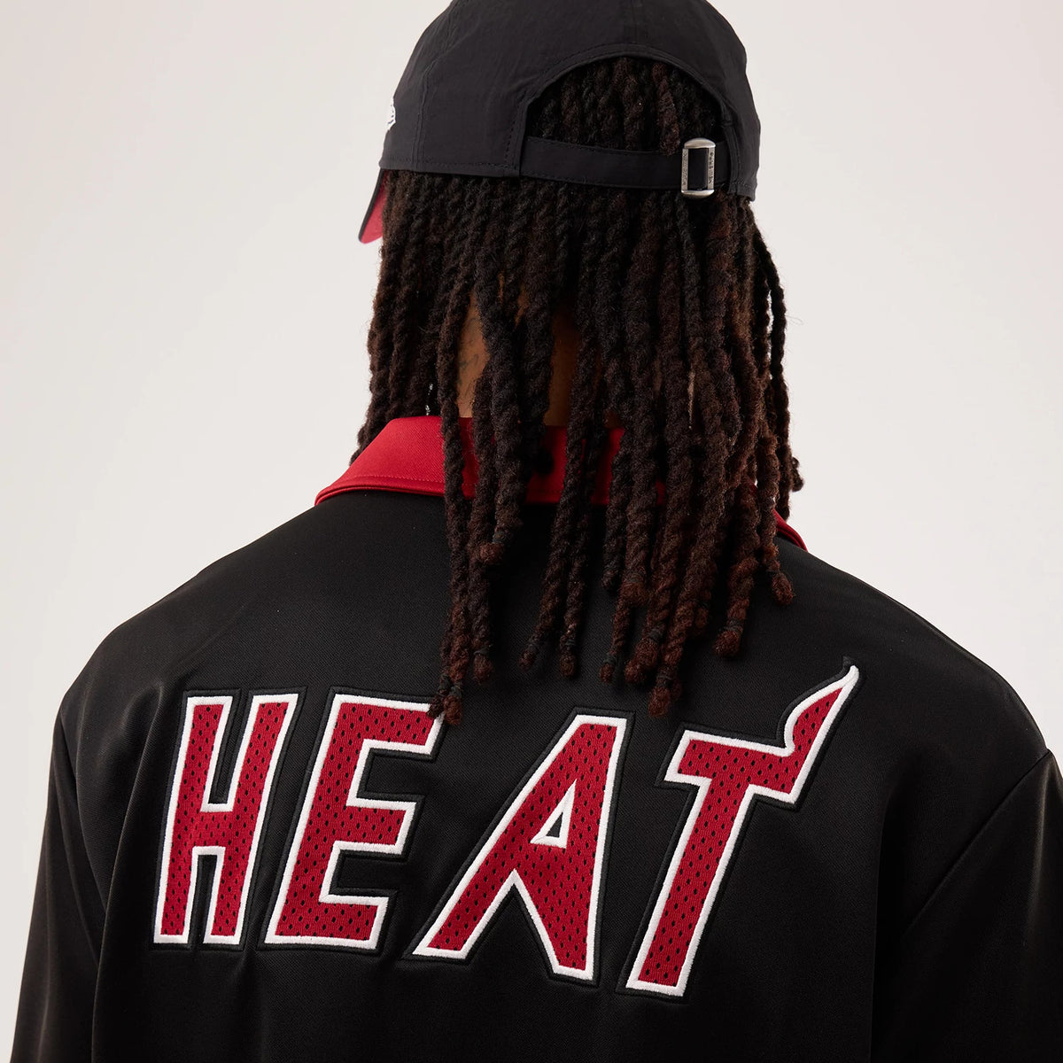 New Era Miami Heat NBA Hardwood Classic Revere Black Short Sleeve Shirt, back view on a model showing the large red and white embroidered HEAT lettering across the upper back, complemented by a black cap with an adjustable strap.