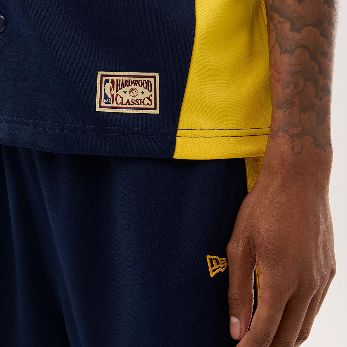 New Era Warriors NBA Hardwood Classic Revere Dark Blue Short Sleeve Shirt, close-up on a model showing the NBA Hardwood Classics patch on the dark blue and contrasting yellow fabric, with the New Era flag logo embroidered on the left side.