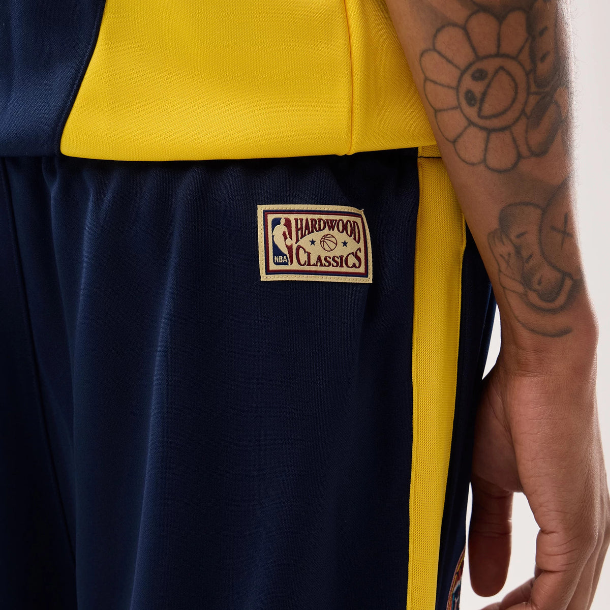 New Era Warriors NBA Hardwood Classic Straight Dark Blue Straight Leg Joggers, side view on a model with tattoos, showing the NBA Hardwood Classics patch on the upper left leg, dark blue fabric, and yellow side stripe detail.