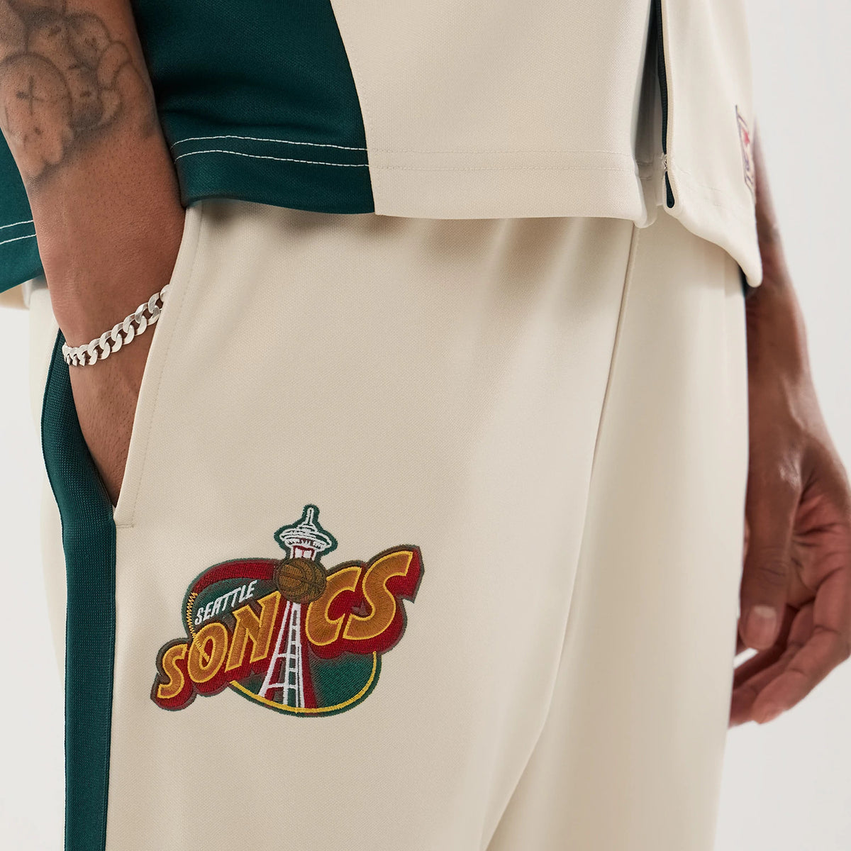 New Era Supersonics NBA Hardwood Classic Beige Straight Leg Joggers, close-up view showing the Seattle Supersonics logo embroidered on the right leg, with green side stripe detail and beige fabric, worn by a model with a chain bracelet.