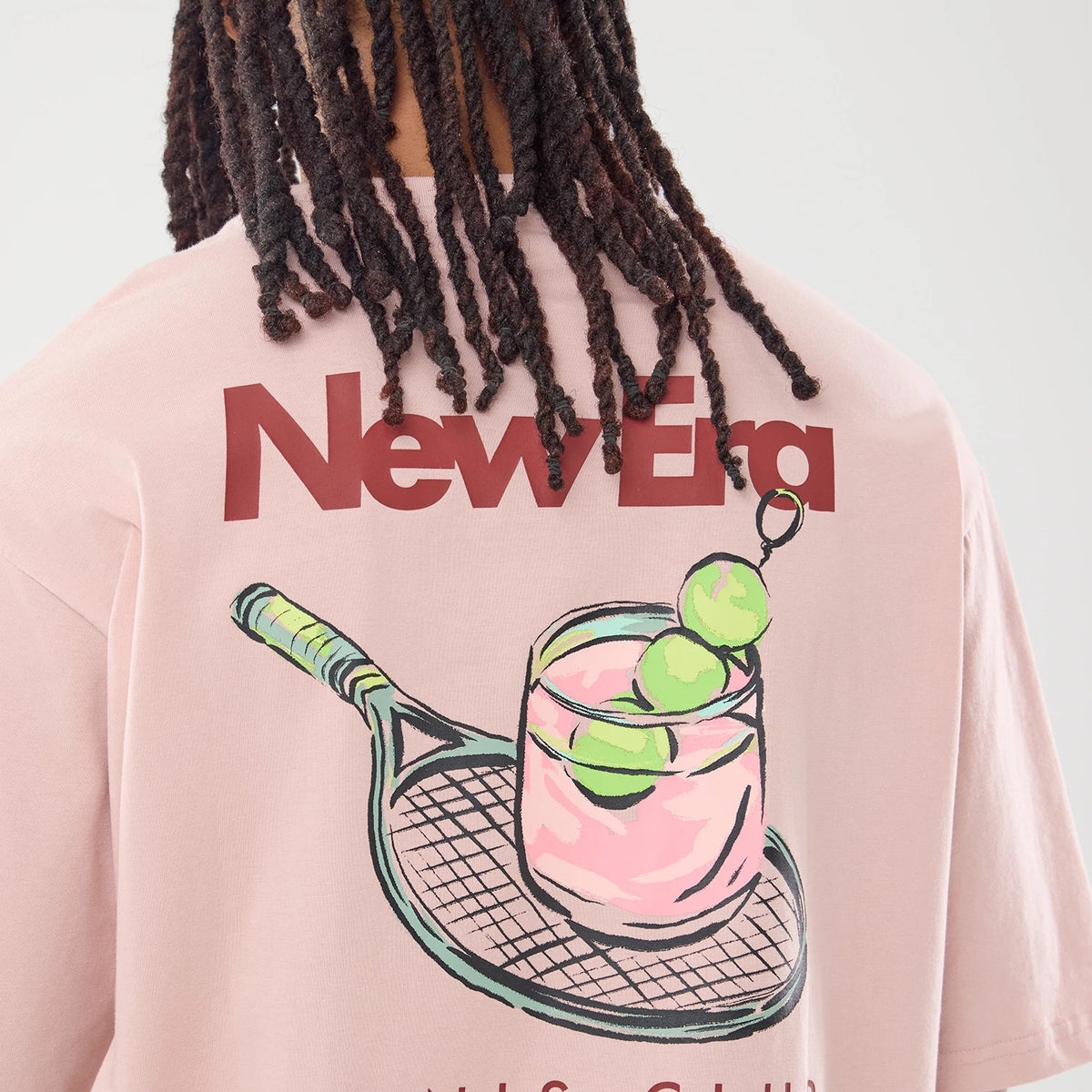 New Era Tennis Graphic Oversized Pastel Pink T-Shirt, back view showing a model wearing the shirt with a large printed graphic of a tennis racket and a glass with green balls, and red New Era lettering prominently across the upper back.