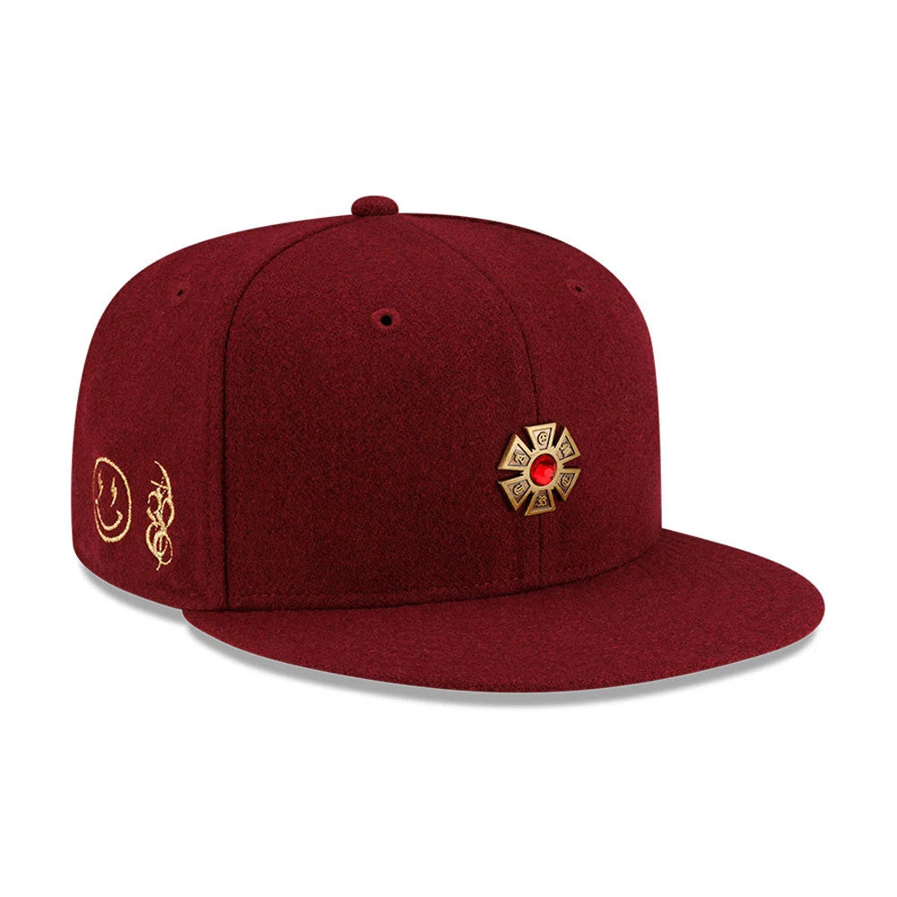 New Era X Omerta Balvin Dark Red 59FIFTY Fitted Cap, front view showing a emblem with a red centrepiece on the right panel, a gold embroidered smiley face and script on the left, and a flat dark red visor.