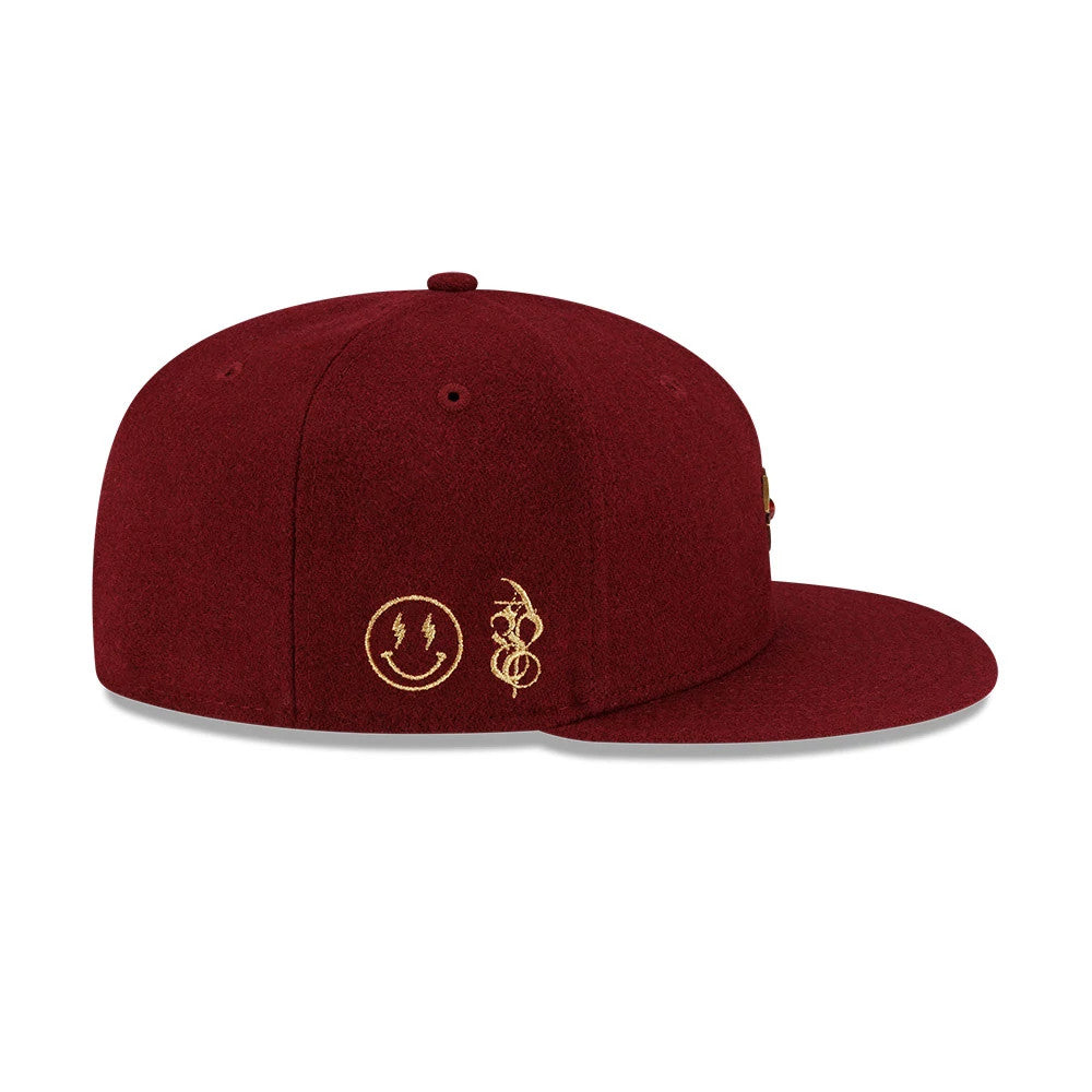 New Era X Omerta Balvin Dark Red 59FIFTY Fitted Cap, side view showing a structured dark red crown with embroidered gold symbols including a smiley face and swooping design, set above the gently curved visor.