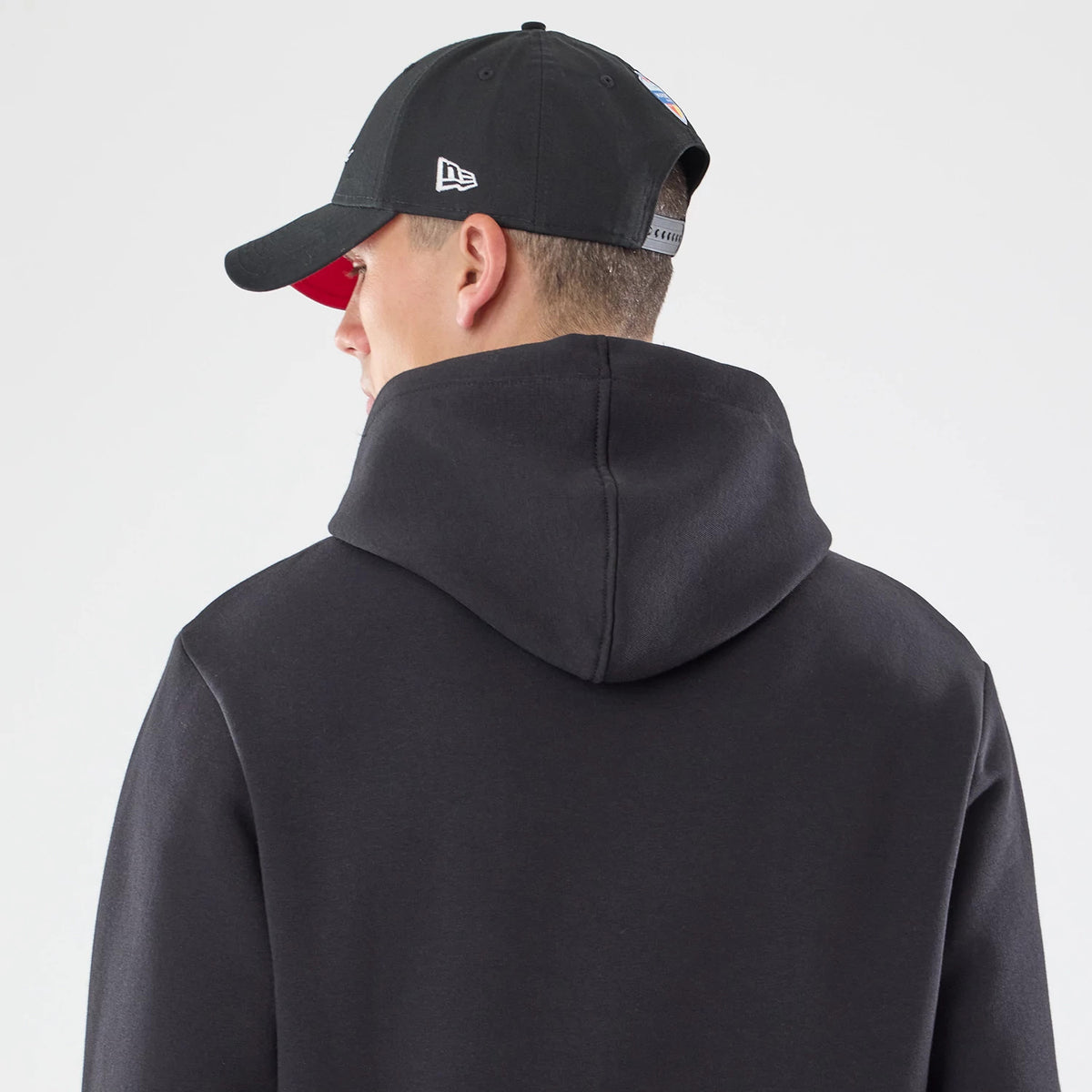 New Era Atlanta Falcons NFL Patch Black Pullover Hoodie, back view on a model wearing a black cap, showing the hood's seam structure and plain surface, with the cap featuring an embroidered New Era logo on the side and adjustable strap at the back.