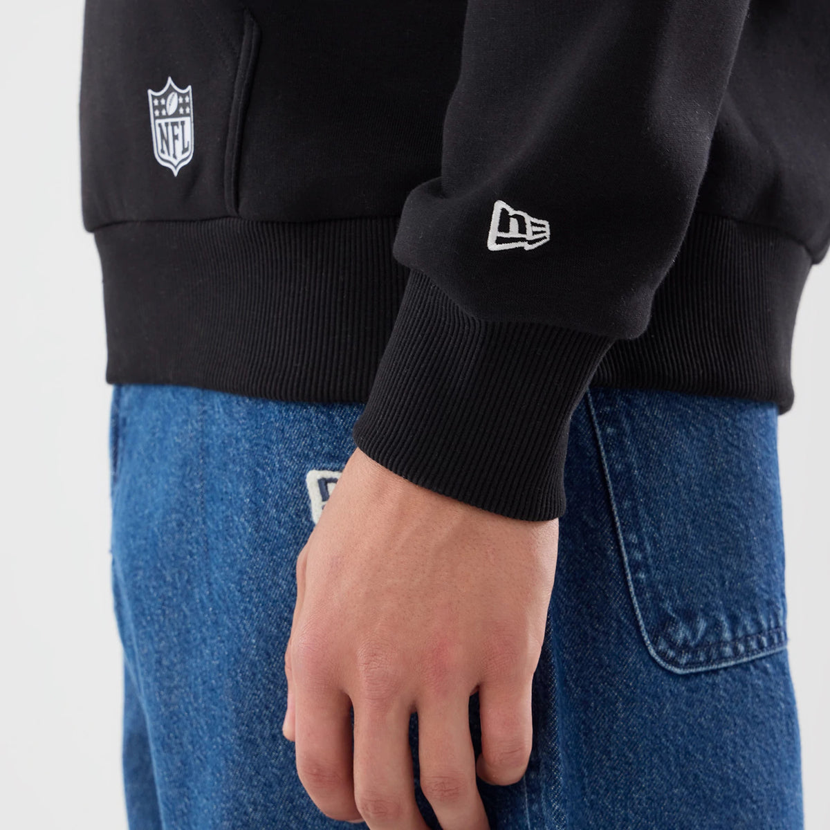 New Era New Orleans Saints NFL Patch Black Pullover Hoodie, side view showing the black ribbed cuff with a white embroidered New Era logo, an embroidered NFL patch on the pocket and the hoodie styled casually with blue jeans.