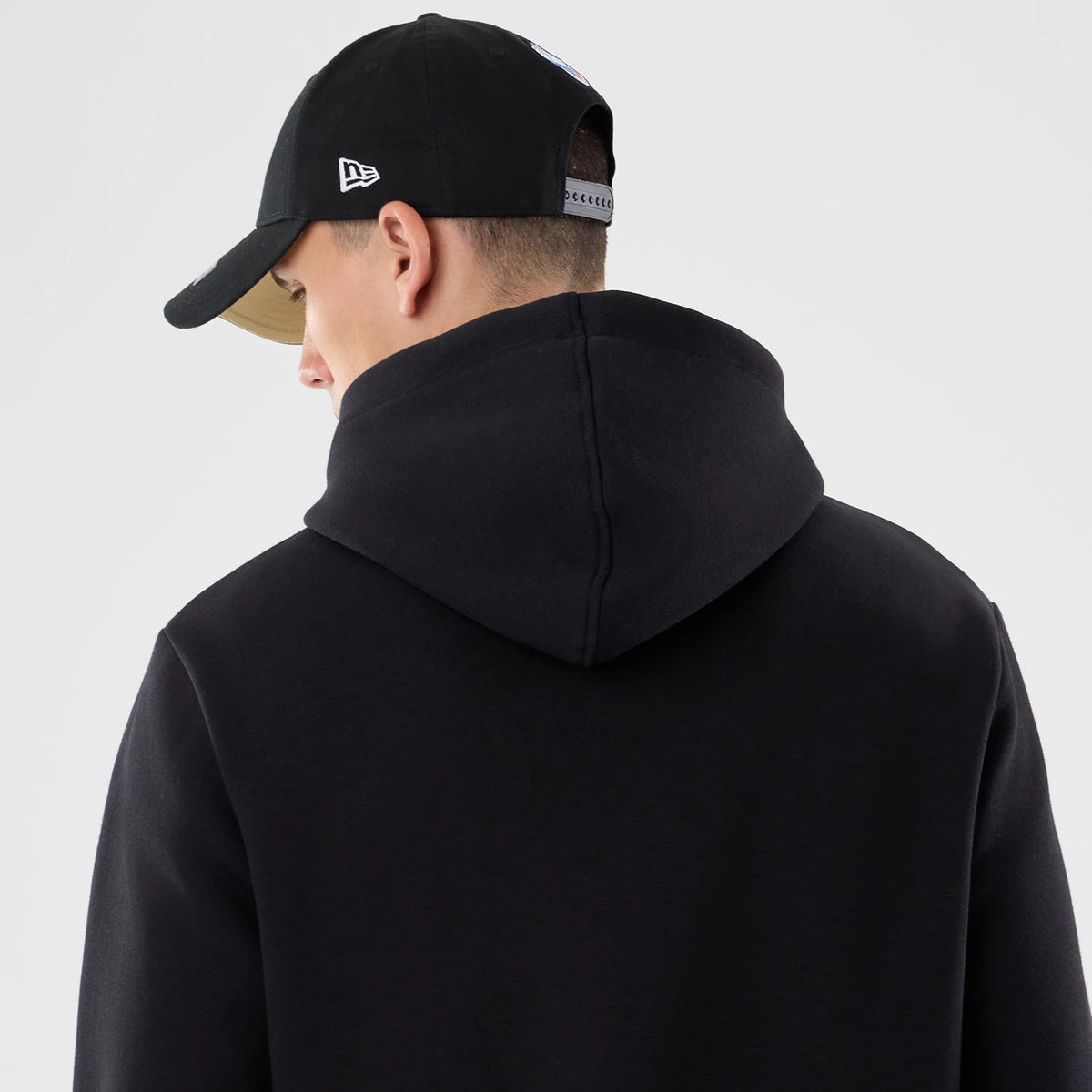 New Era New Orleans Saints NFL Patch Black Pullover Hoodie, back view on a model showing the plain black hood and upper back, paired with a black cap featuring an embroidered New Era logo on the side panel.