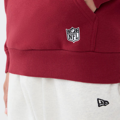 New Era Washington Commanders NFL Patch Dark Red Pullover Hoodie, close-up view showing a white embroidered NFL shield logo on the front pocket, a ribbed hem, and the New Era logo on matching light grey trousers worn by the model.