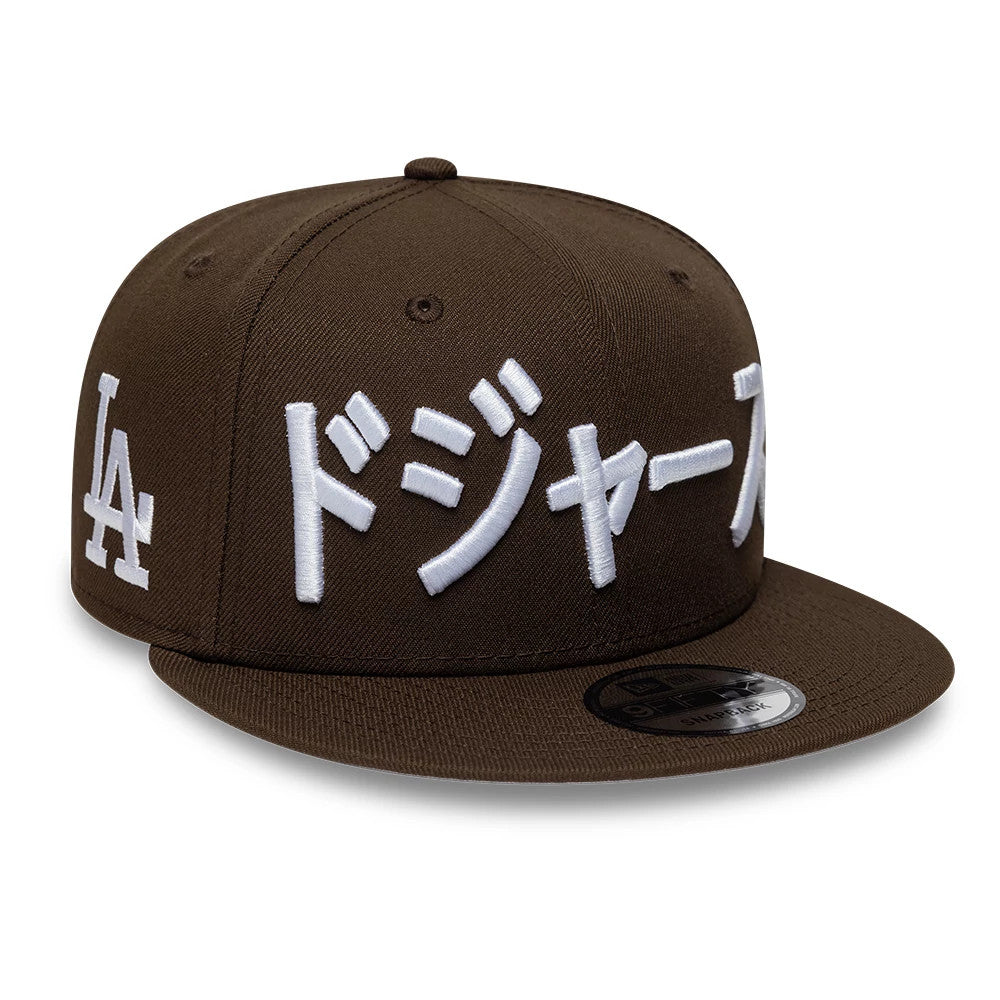 LA Dodgers MLB Kanji Dark Brown New Era 9FIFTY Snapback Adjustable Cap, three-quarter view showing white raised embroidered Kanji script across the front panels above the flat dark brown visor, with a 9FIFTY size sticker and LA logo on the side.