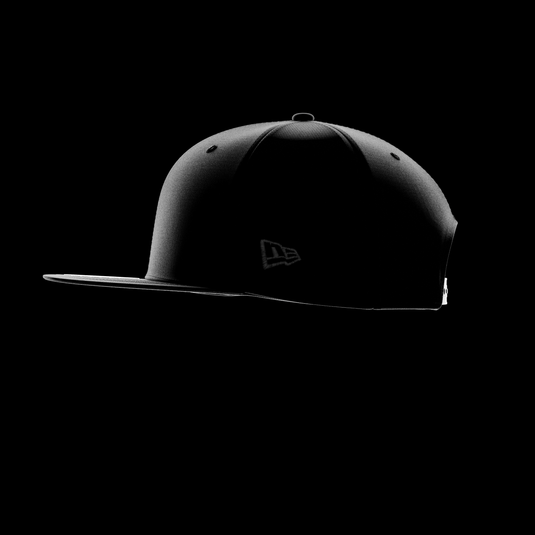 A black 9FIFTY cap shown in side profile against a solid black background, featuring a subtle tonal New Era Flag on the side and a structured flat brim.