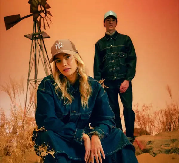 A woman wearing a dark blue denim jacket and matching wide-leg jeans sits on a sandy ridge in a desert landscape, while a man in a dark green denim jacket and pants stands behind her. Both are wearing New York Yankees A-Frame Caps.