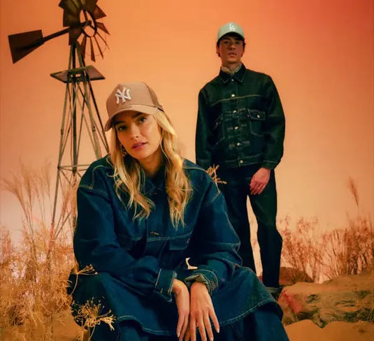 A woman wearing a dark blue denim jacket and matching wide-leg jeans sits on a sandy ridge in a desert landscape, while a man in a dark green denim jacket and pants stands behind her. Both are wearing New York Yankees A-Frame Caps.