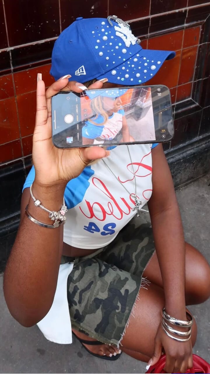 Girl kneeling down wearing a blue and white New York Yankees cap and a camp skirt, holding her phone in her hand showing the screen.