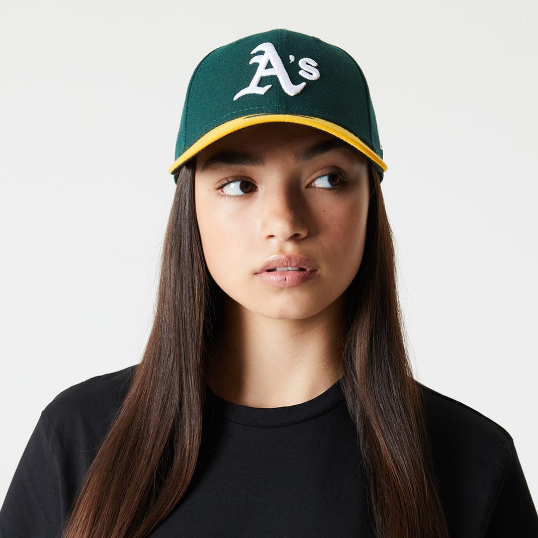 A girl wearing a green Oakland Athletics cap with a yellow brim and white embroidered 'A's' logo, paired with a black crewneck shirt against a light gray background.