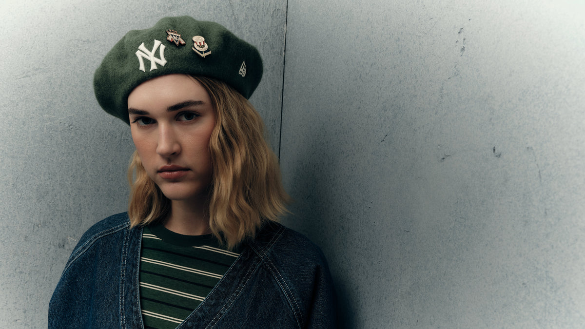 A person with shoulder-length blonde hair leans against a textured gray wall, wearing a dark green New York Yankees beret, a blue denim jacket with a white 'NE' graphic, and a green and black striped t-shirt.