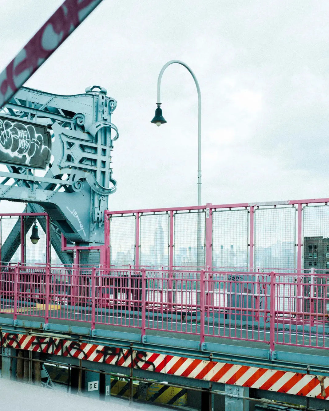 Part of a New York bridge with a pink railing and a streetlight against a grey sky.
