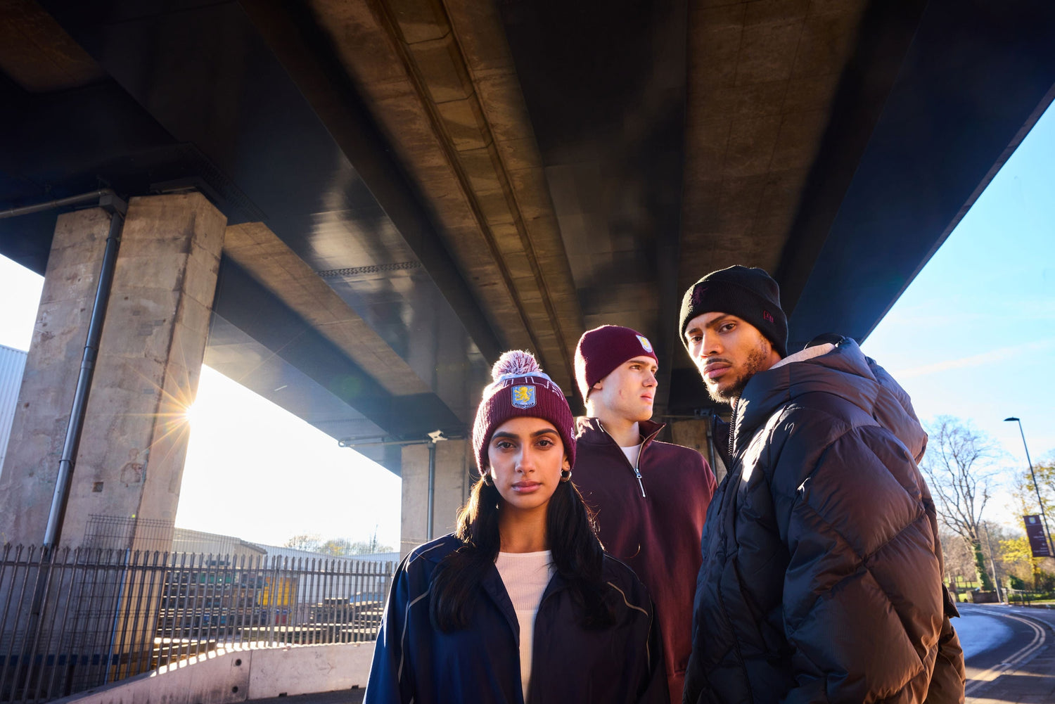 Three people standing under a bridge with a road and trees in the background wearing new era aston villa beanies