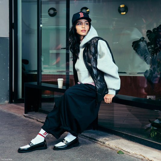 A woman sits on a black bench outside a storefront, wearing a black Boston Red cap, a white hoodie with a black lace-up vest, loose black pants, white socks, and black-and-white loafers.