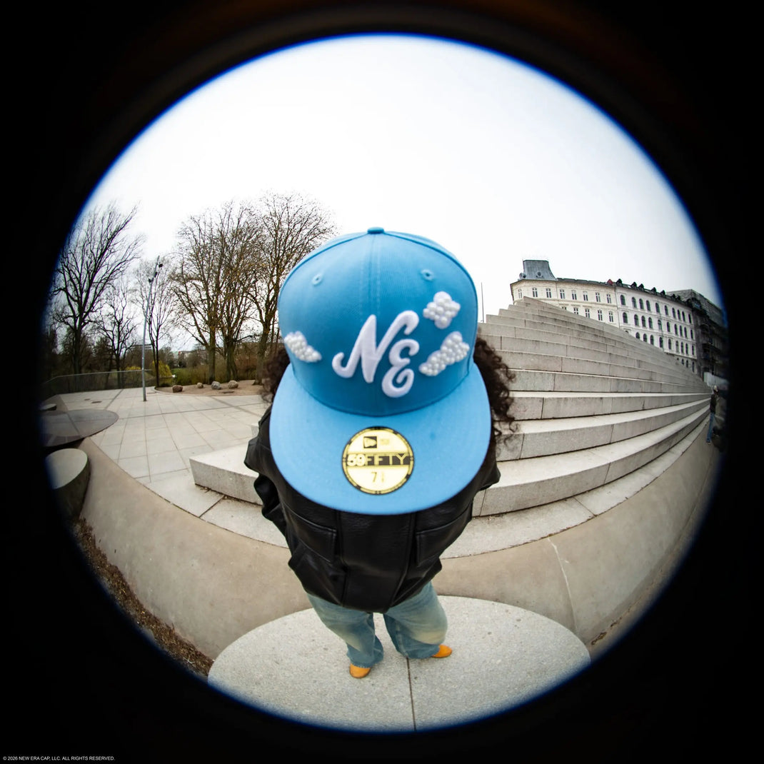 Person wearing a blue new era 59fifty cap with white text standing on steps outdoors.