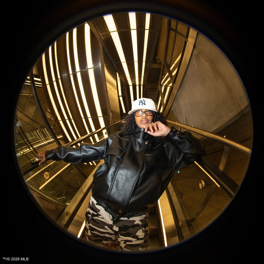 Person wearing a white 59fifty cap, black leather jacket, and camouflage trousers in a reflective, circular mirror.