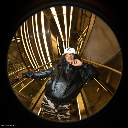 Person wearing a white 59fifty cap, black leather jacket, and camouflage trousers in a reflective, circular mirror.
