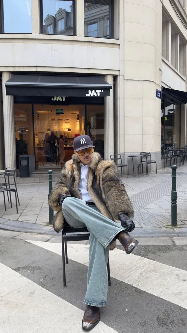 A man sitting on a chair on in the road on a zebra crossing wearing a navy 59FIFTY New York Yankees cap, yellow tinted glasses, fur coat, blue jeans and brown boots.