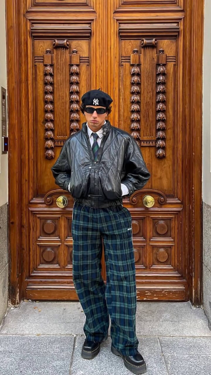A man standing in front of a large brown door wearing a black New Era New York Yankee beret, black sunglasses, shirt and tie, a black leather jacket with blue and green chequered trousers and black platform shoes.