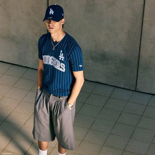 A man wearing a navy blue pinstriped Los Angeles Dodgers t-shirt, gray shorts, blue LA cap, and white socks, standing in a tiled indoor space with a concrete wall background.