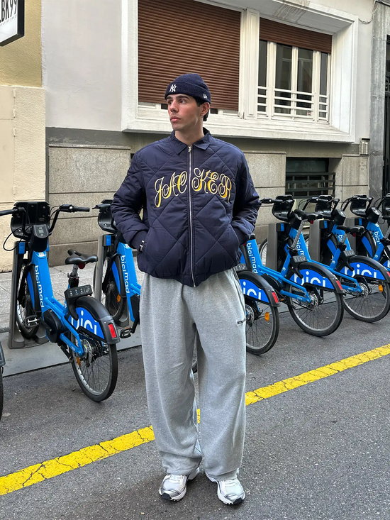 Slide 4:Person wearing a navy bomber jacket with yellow text and new era beanie, standing in front of blue bicycles on a street.
