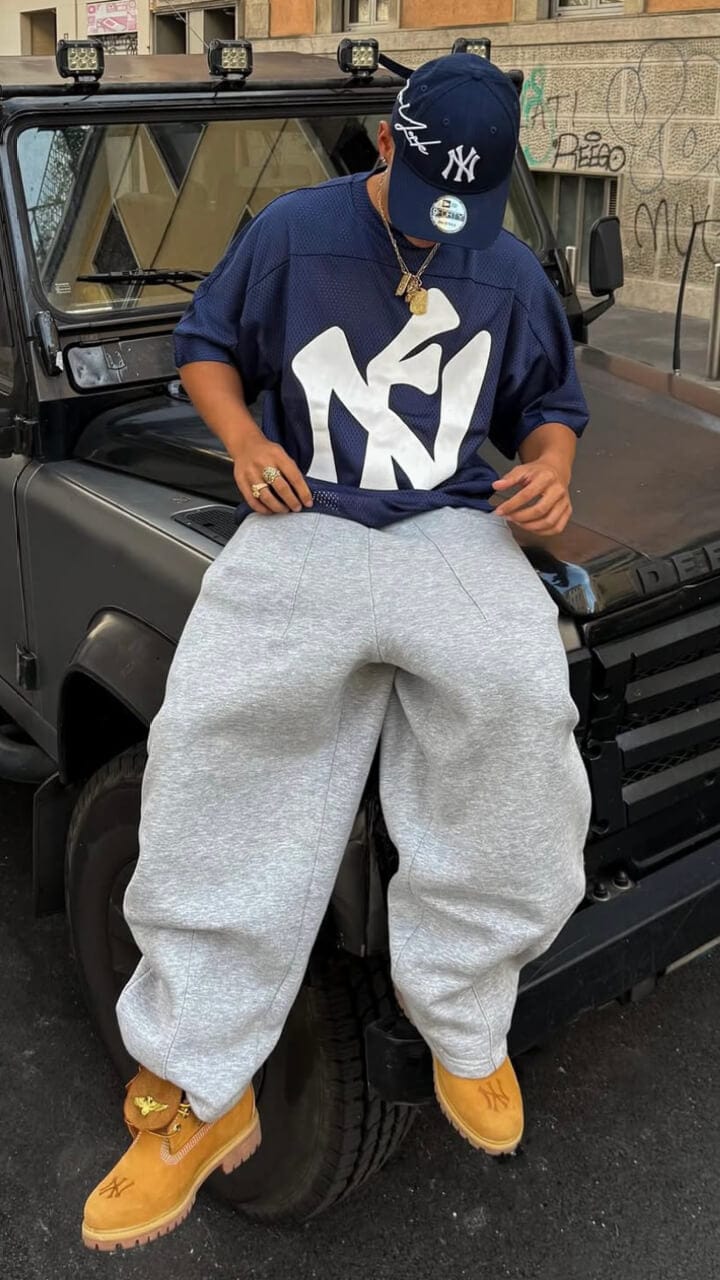 A man sitting on a black Jeep wearing a a navy New York Yankees New era cap and t-shirt, grey joggers and nubuck Timberland boots.