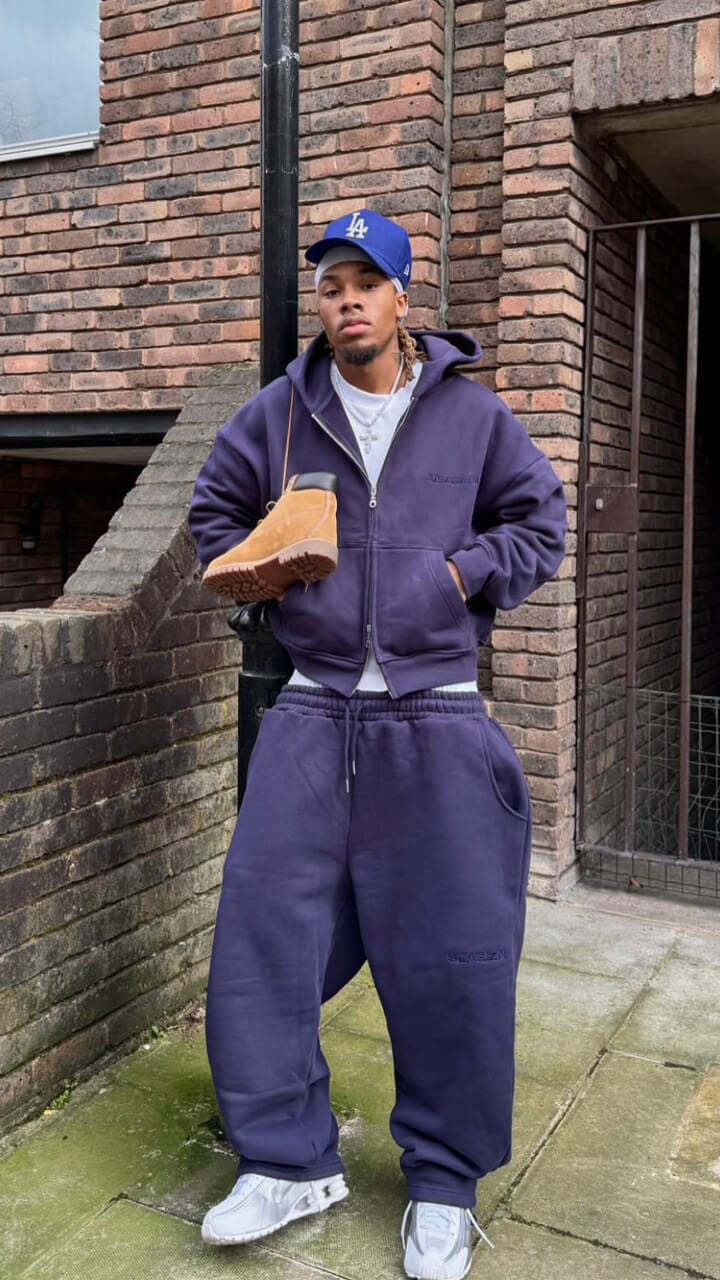 A man leaning on a street lamp wearing a blue LA Dodgers New Era cap, navy tracksuit and white trainers.