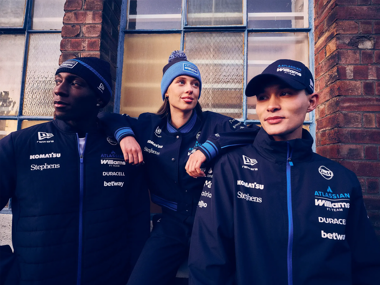 Three individuals wearing matching dark blue williams motorsports jackets and new era hats with various logos in front of a brick wall.