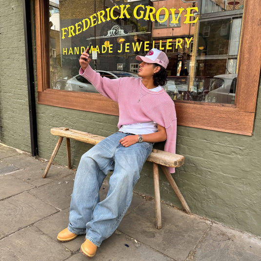Person sitting on a stool wearing a New Era trucker cap in front of 'Frederick Grove Handmade Jewellery' store.