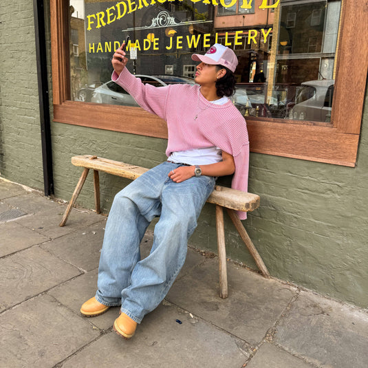 Person taking a selfie on a wooden stool in front of a jewelry store wearing a pink trucker cap.