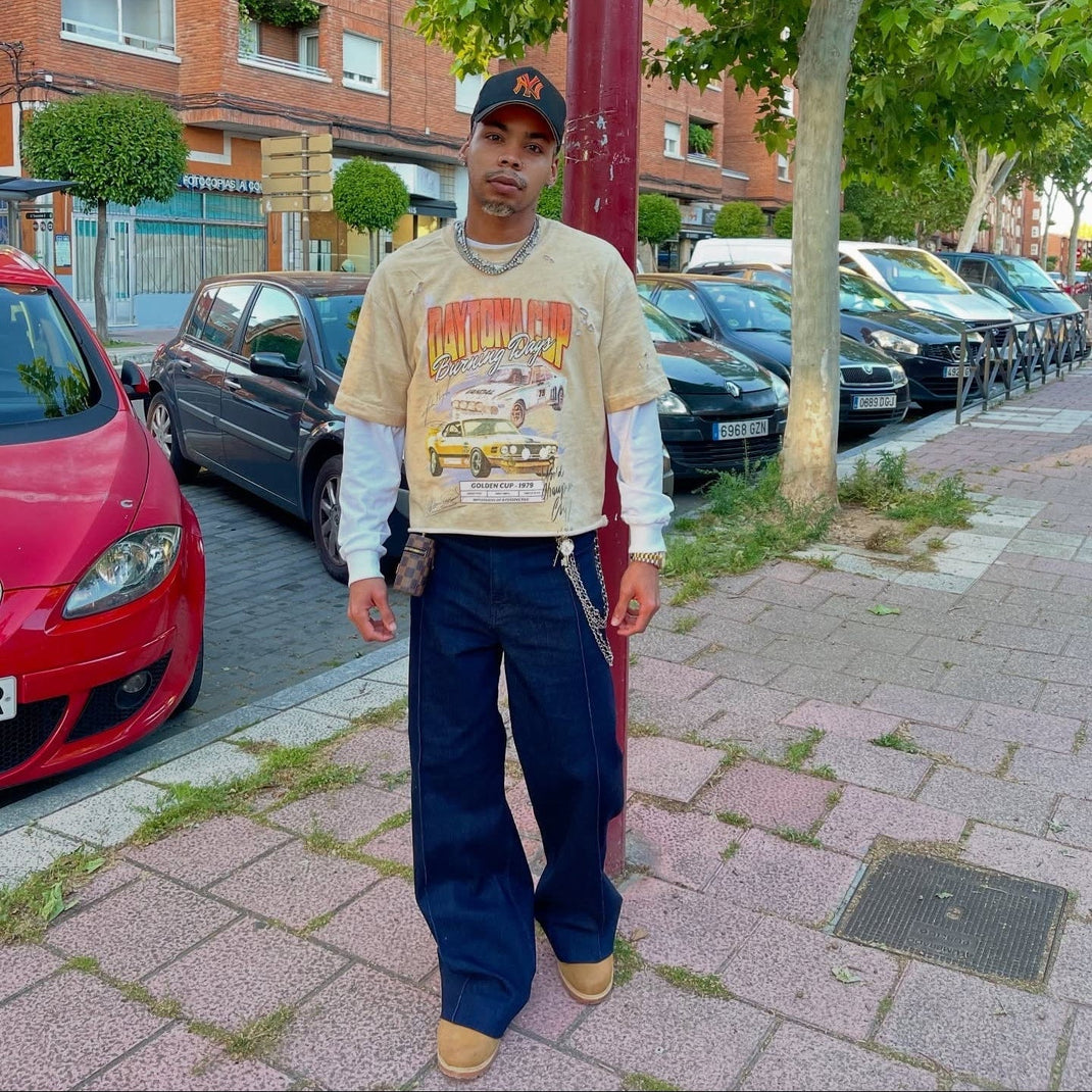 Person wearing a graphic sweatshirt and blue pants standing on a sidewalk with parked cars and buildings in the background.
