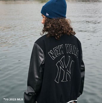 woman on beach wearing a black new york yankees varsity jacket and a navy new era beanie.